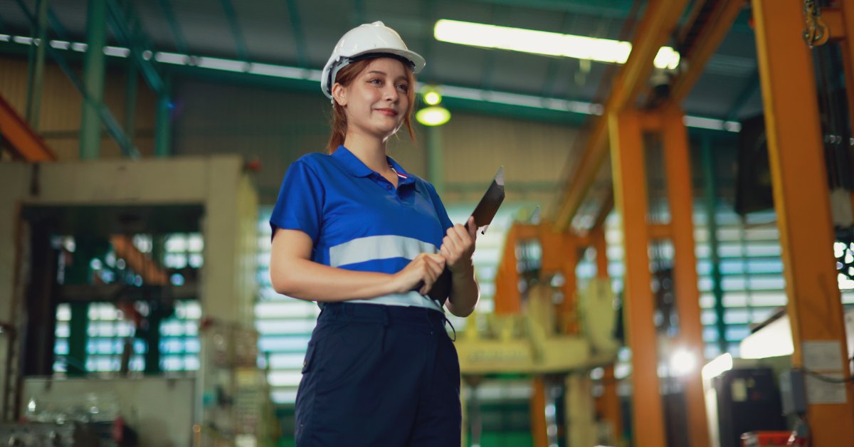 Worker in a hard hat and blue safety uniform holds a tablet on a factory floor surrounded by large equipment.