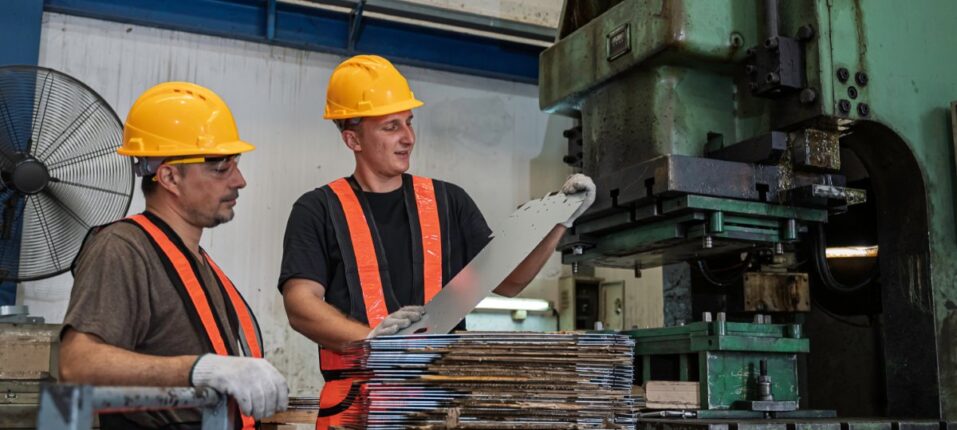 Two workers wearing safety gear handle stacked metal sheets while operating a large green machine in a factory.