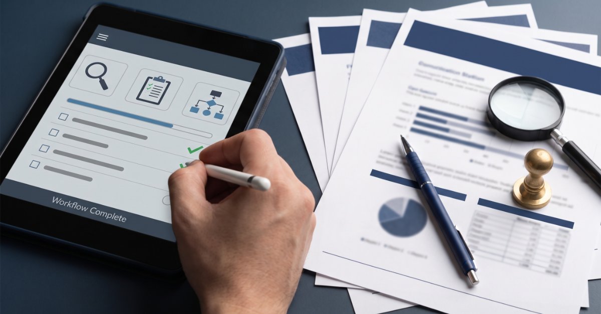 Hand checks items on a digital workflow displayed on a tablet beside printed reports, a pen, and magnifying glass.