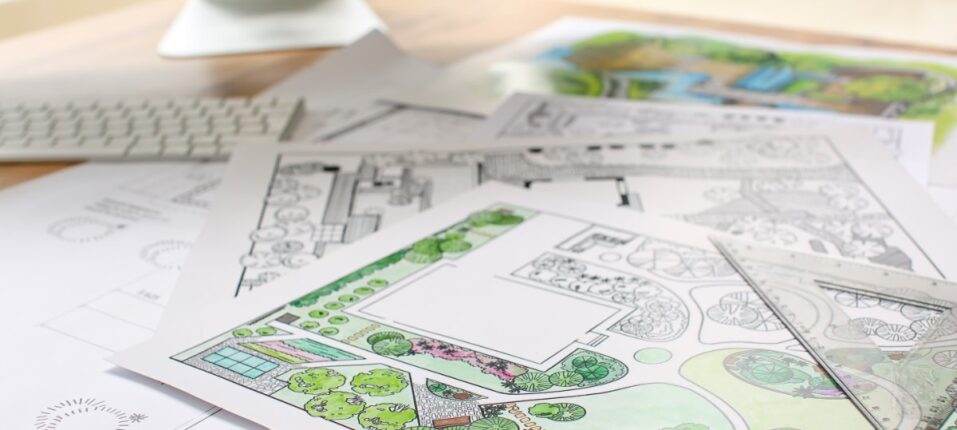 Landscape design plans with colorful garden layouts and planting details spread across a desk beside a computer.