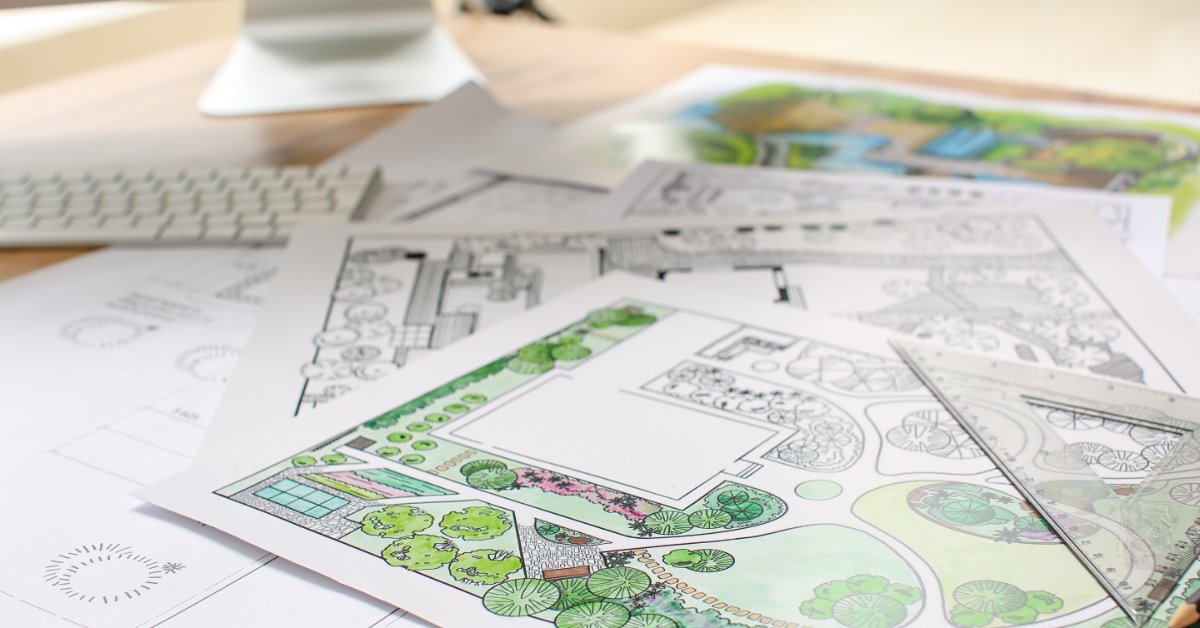 Landscape design plans with colorful garden layouts and planting details spread across a desk beside a computer.