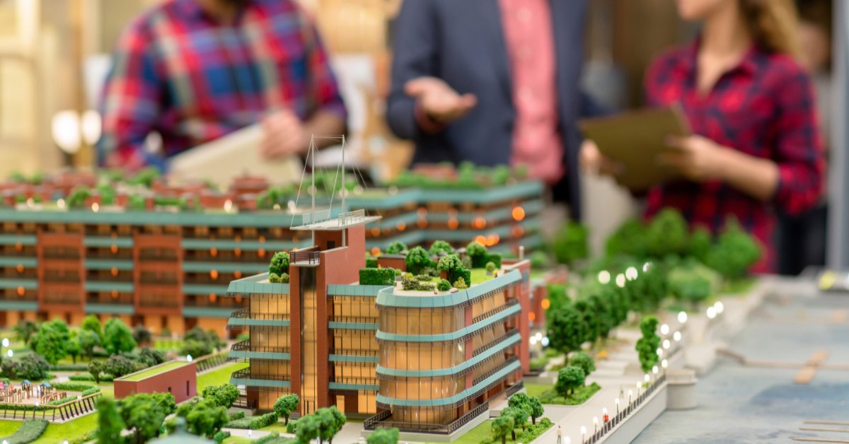 Three people review plans behind a detailed architectural model featuring modern buildings, trees, and a waterfront.