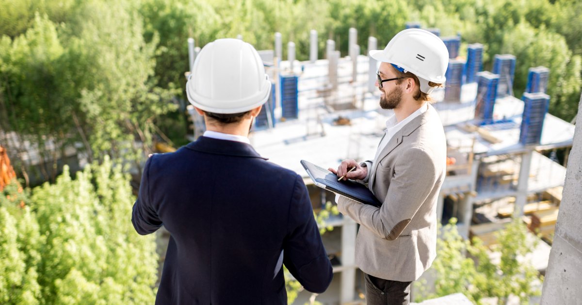 Two men wearing hard hats survey a construction site from above, with one holding a tablet and stylus.